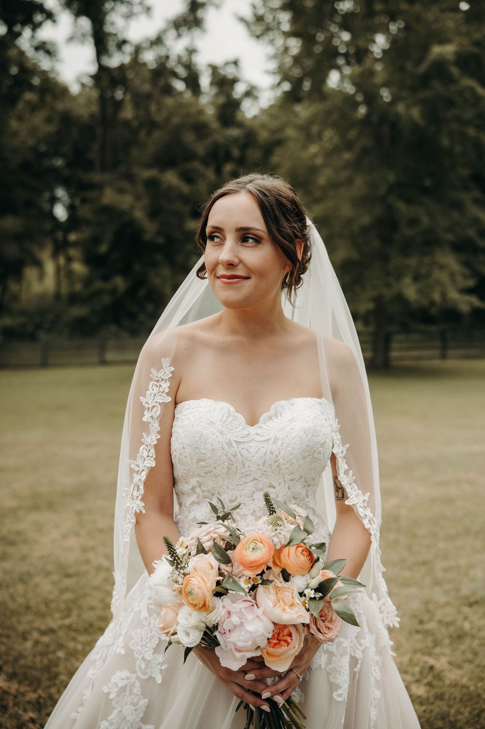 Bride in strapless lace gown with cathedral veil holding peach and blush floral bouquet outdoors.