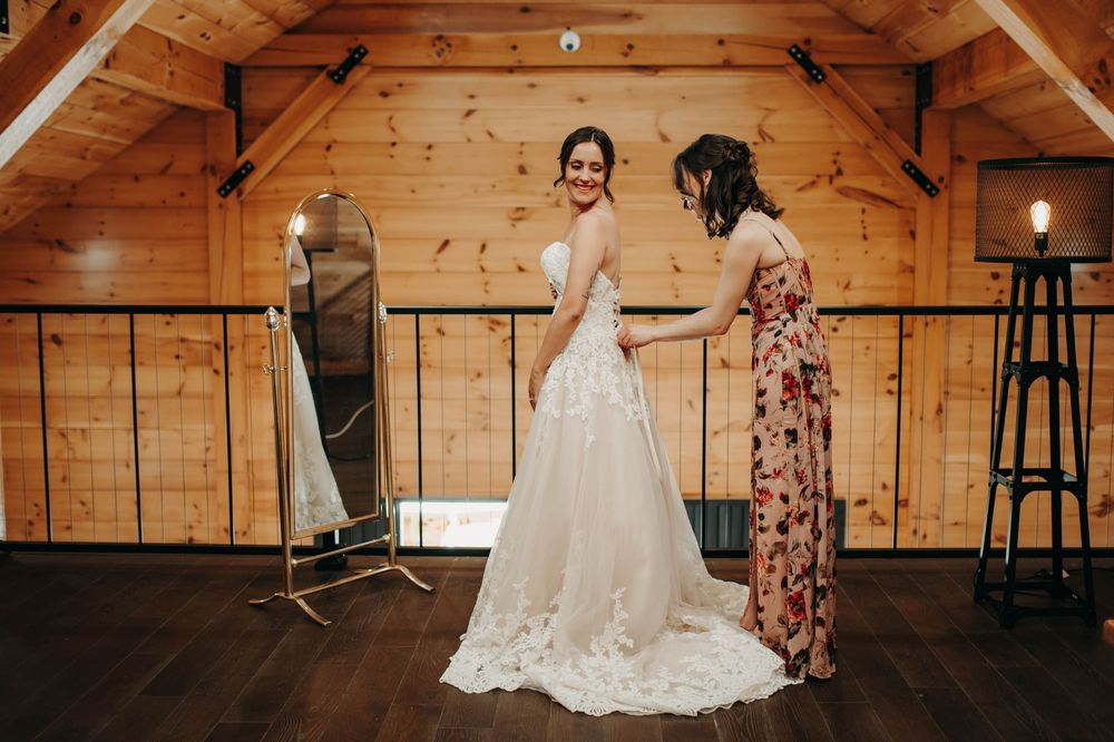 Bridesmaid in floral dress helps bride in lace gown in a wooden barn bridal suite.