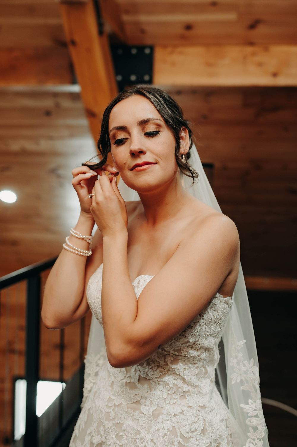 Bride in lace strapless gown putting on earrings inside a rustic wooden venue.