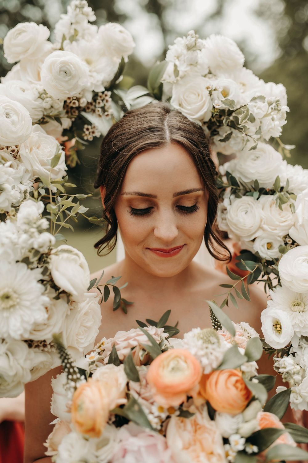 Bride with eyes closed holding peach bouquet framed by white floral arch outdoors.