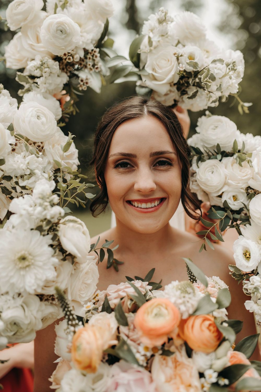 Smiling bride framed by a floral arch of white and peach blooms on her wedding day.