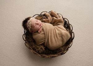 A newborn baby lies peacefully in a brown woven basket against a neutral beige background.