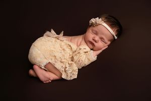 Sleeping newborn baby wearing lace outfit and headband on dark background.