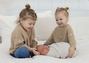 Two young children in matching beige sweaters sit on white bed with newborn baby.