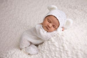 Sleeping newborn baby wearing white knit clothing lies peacefully on soft white textured blanket.