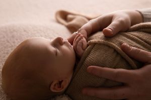 Gentle hands caress a sleeping baby wrapped in a brown knit sweater in soft natural lighting.