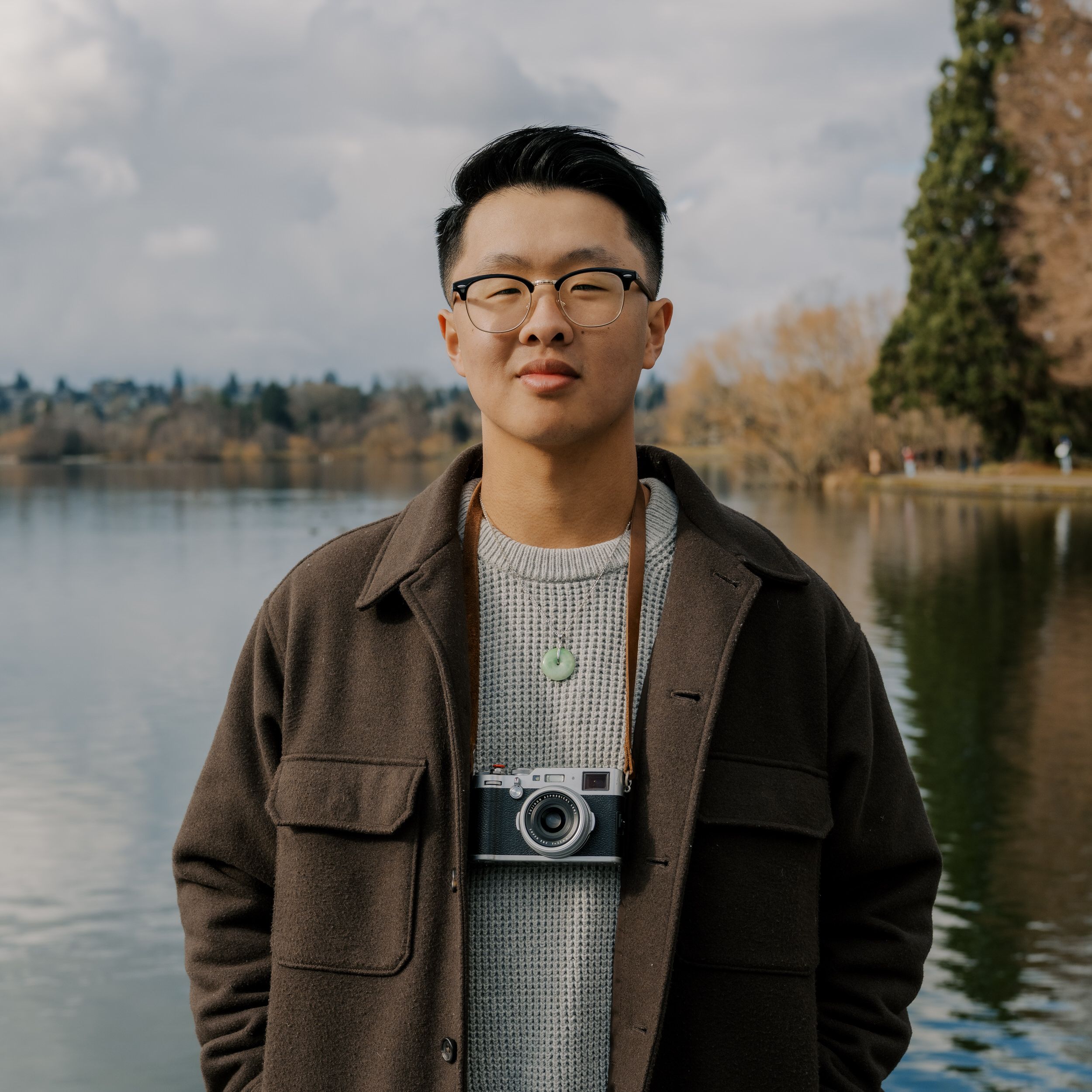A photographer in a brown jacket and gray sweater poses by a serene lake with trees in the background.