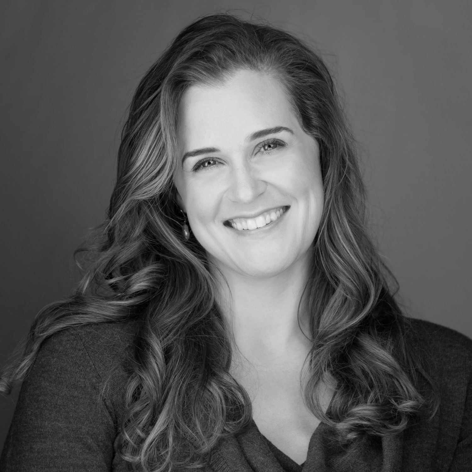 Black and white professional headshot of a woman with long wavy hair and a warm smile wearing dark clothing.