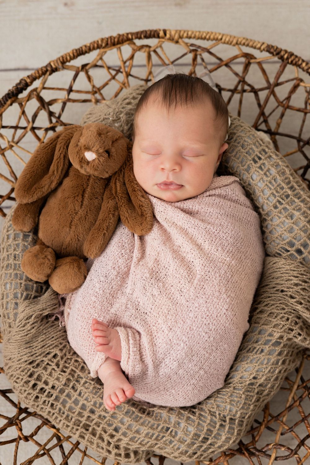 Sleeping newborn wrapped in pink blanket lies in woven basket with teddy bear.