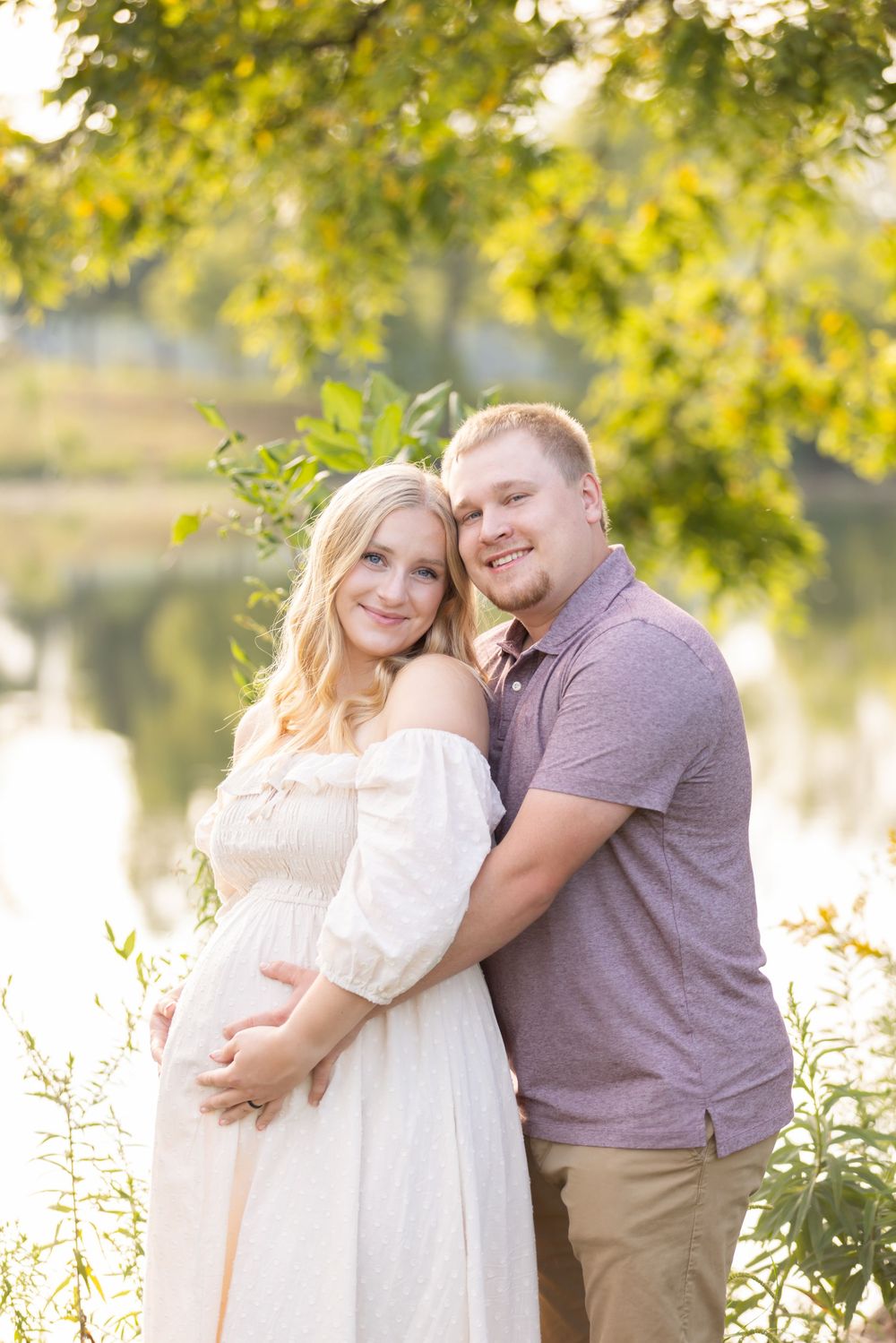 Couple in casual attire embracing outdoors with sunlit trees in background.