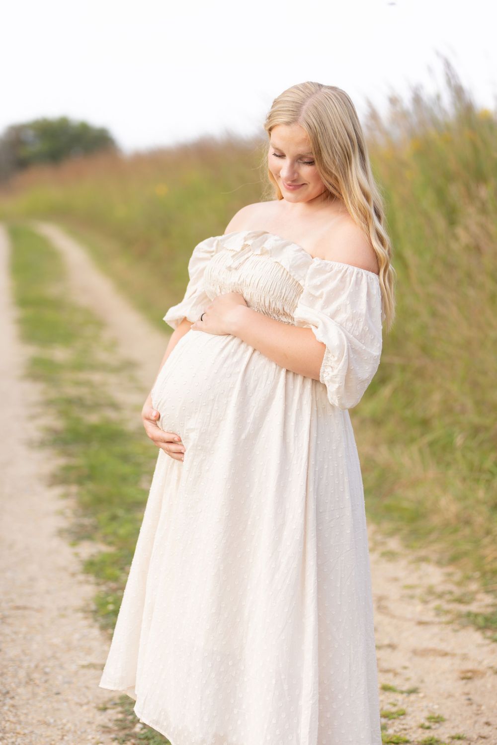 Woman in flowing white dress standing on dirt path in natural setting.