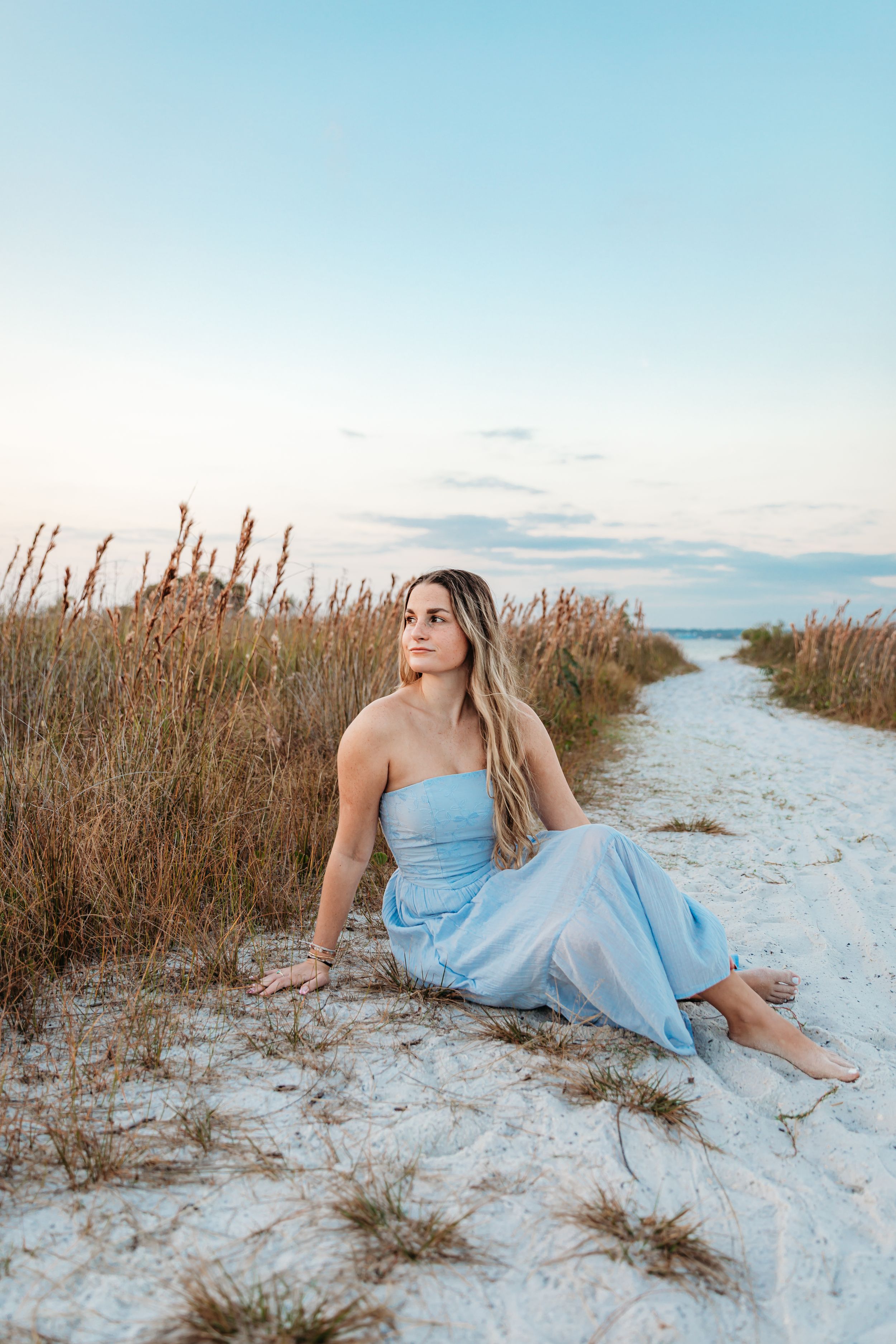 A light blue dress flows in the sea breeze as someone poses on a sandy beach path through tall grass at sunset.