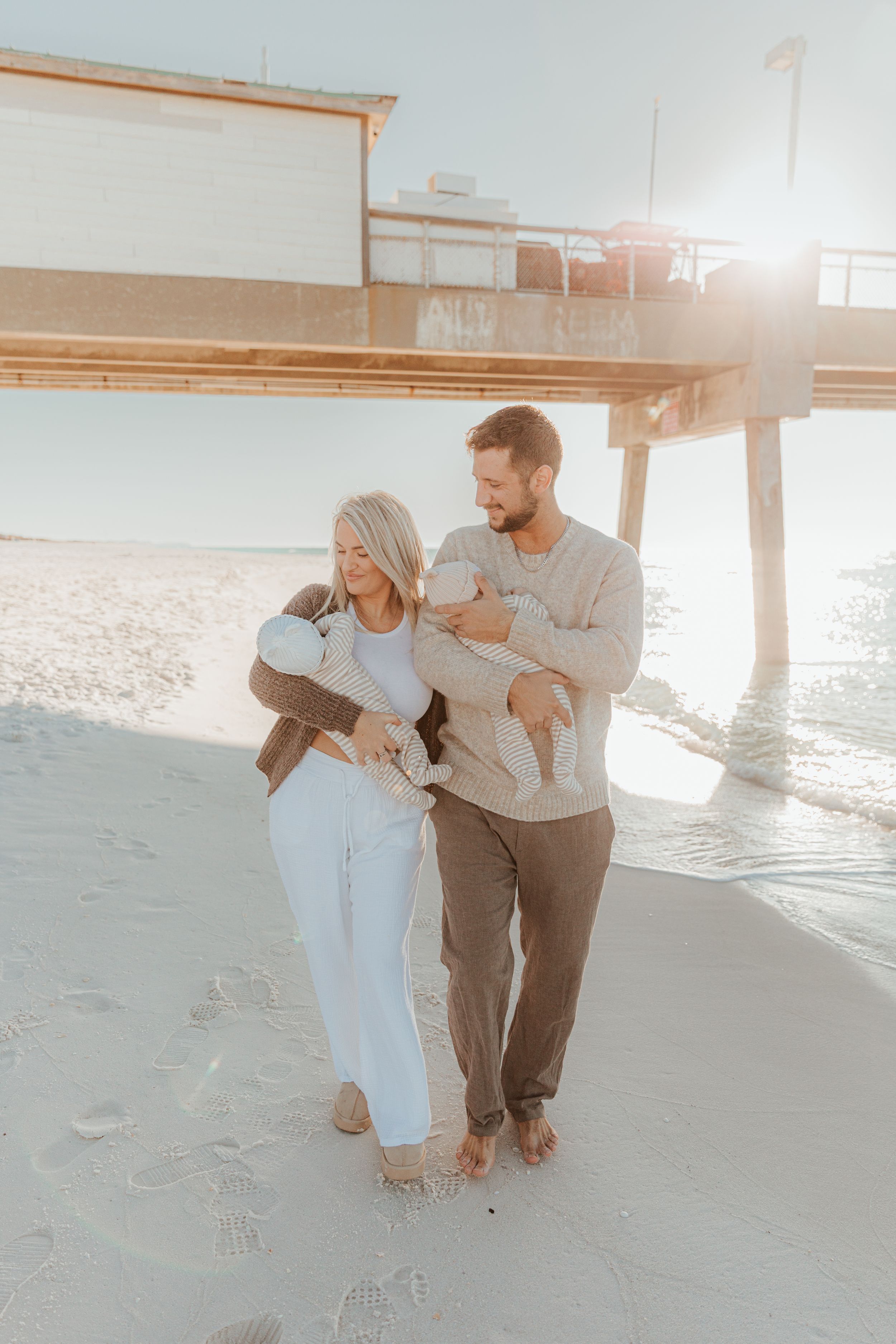A couple holding infants walks barefoot on the beach under a pier during sunset with soft dreamy lighting.