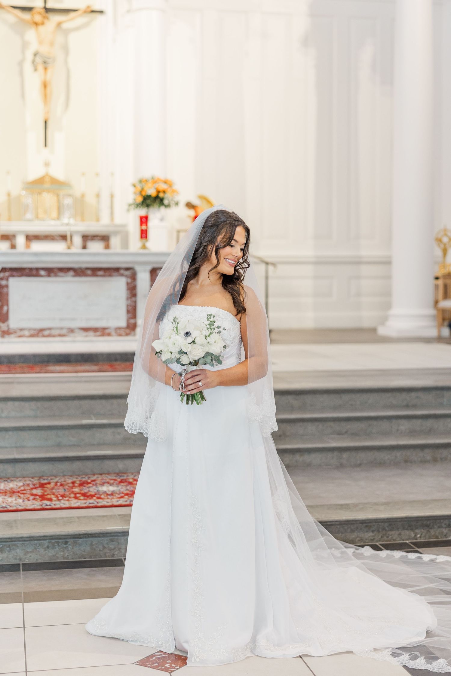 A bride in a flowing white wedding gown and veil stands gracefully in a bright church interior with ornate details.