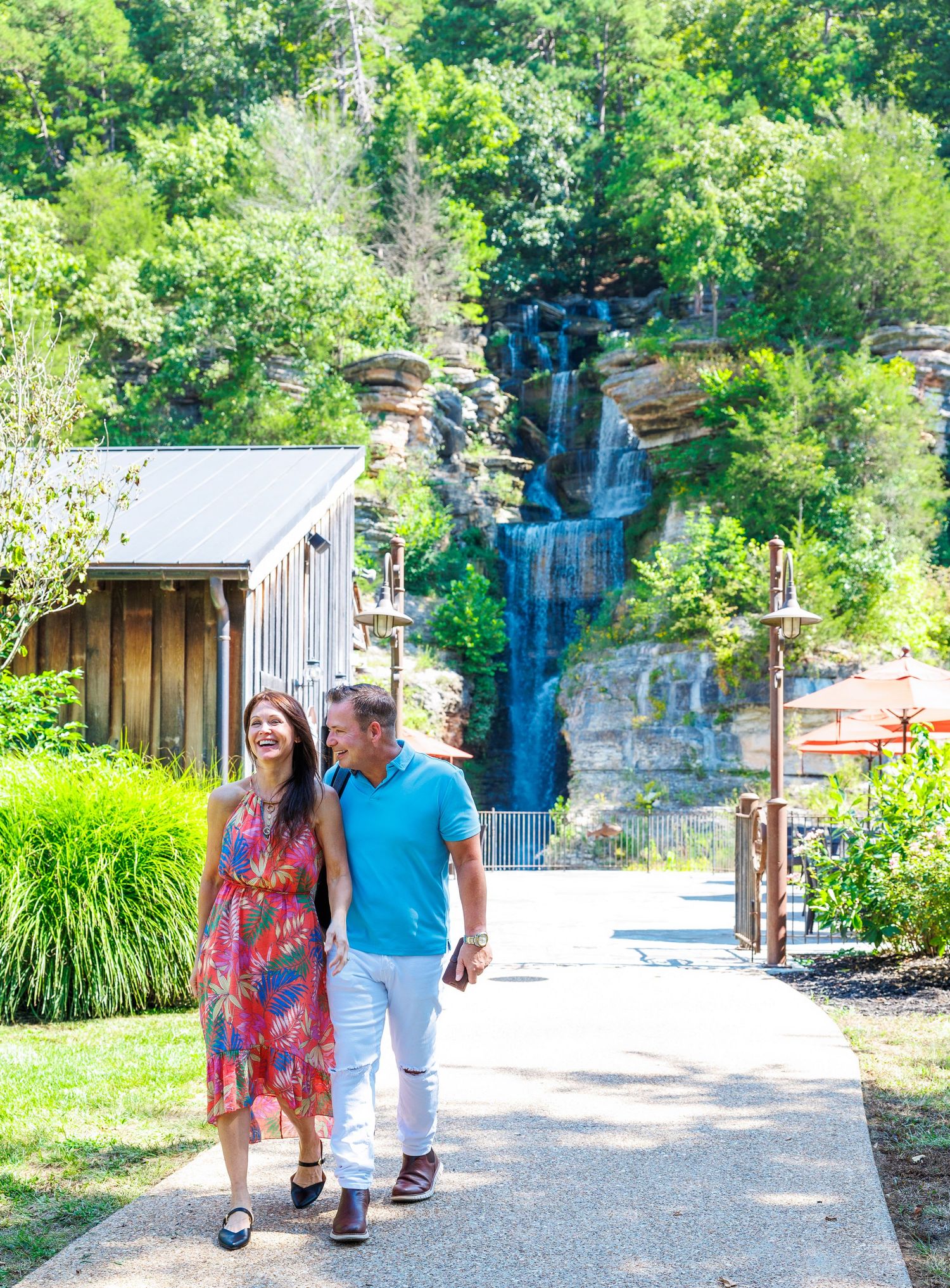 A couple walks along a garden path with a cascading waterfall and lush greenery in the background at a scenic park.