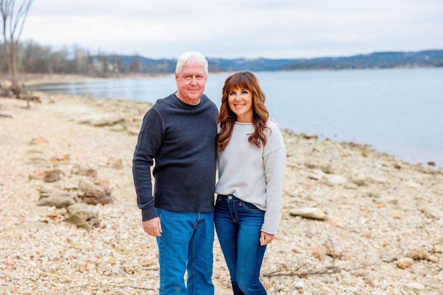 Two people stand together on a rocky beach shore with a calm lake and distant hills in the background at Table Rock Lake.