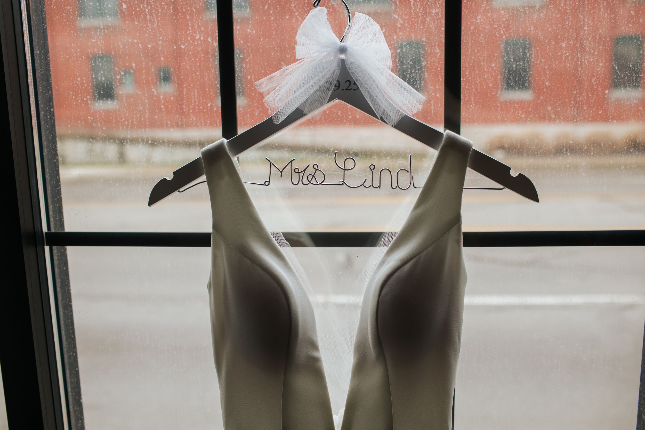 Elegant wedding dress displayed on a hanger against a brick wall with natural window lighting.