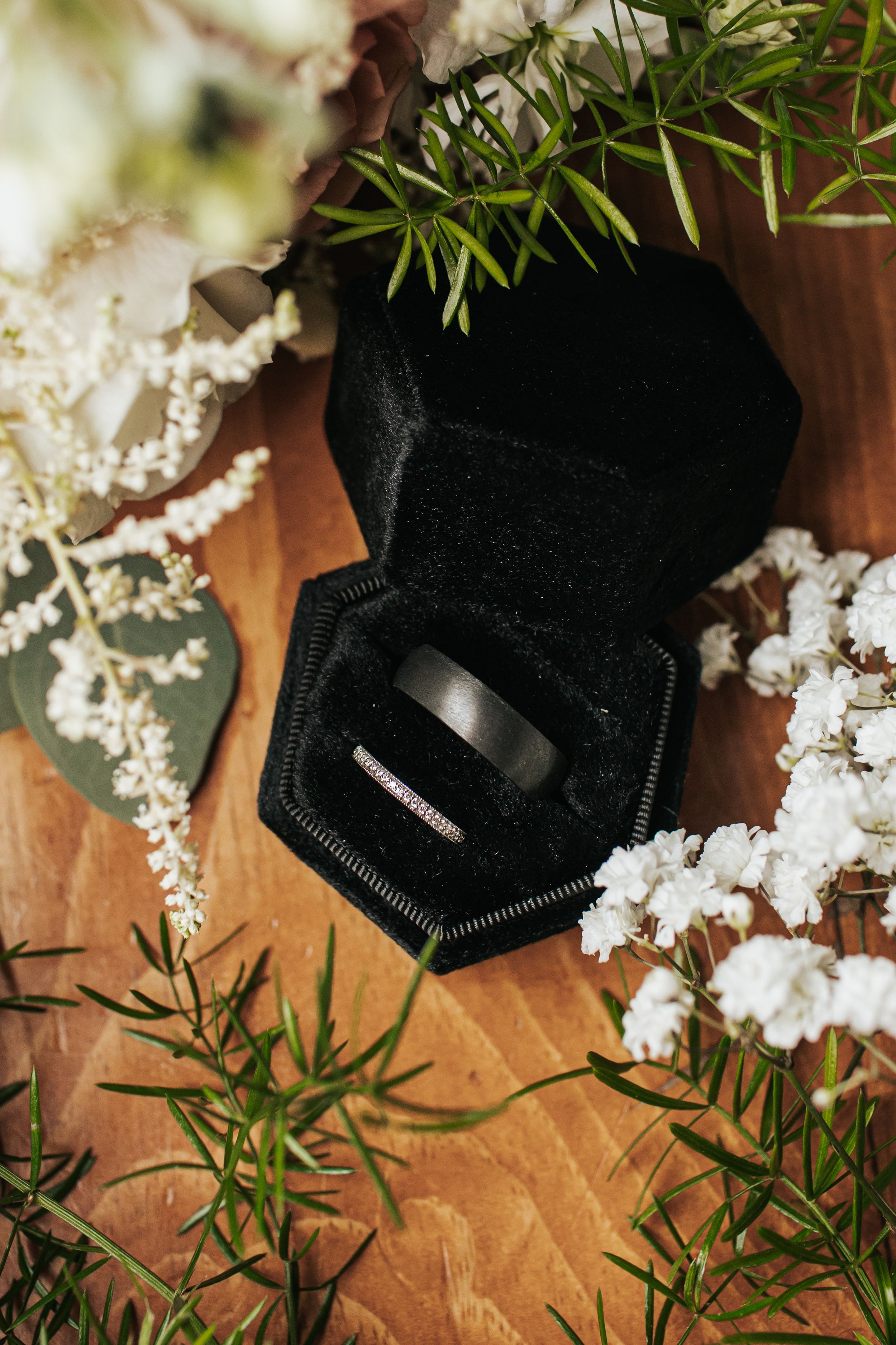 Black wedding band in velvet ring box surrounded by white flowers and greenery on wooden surface.