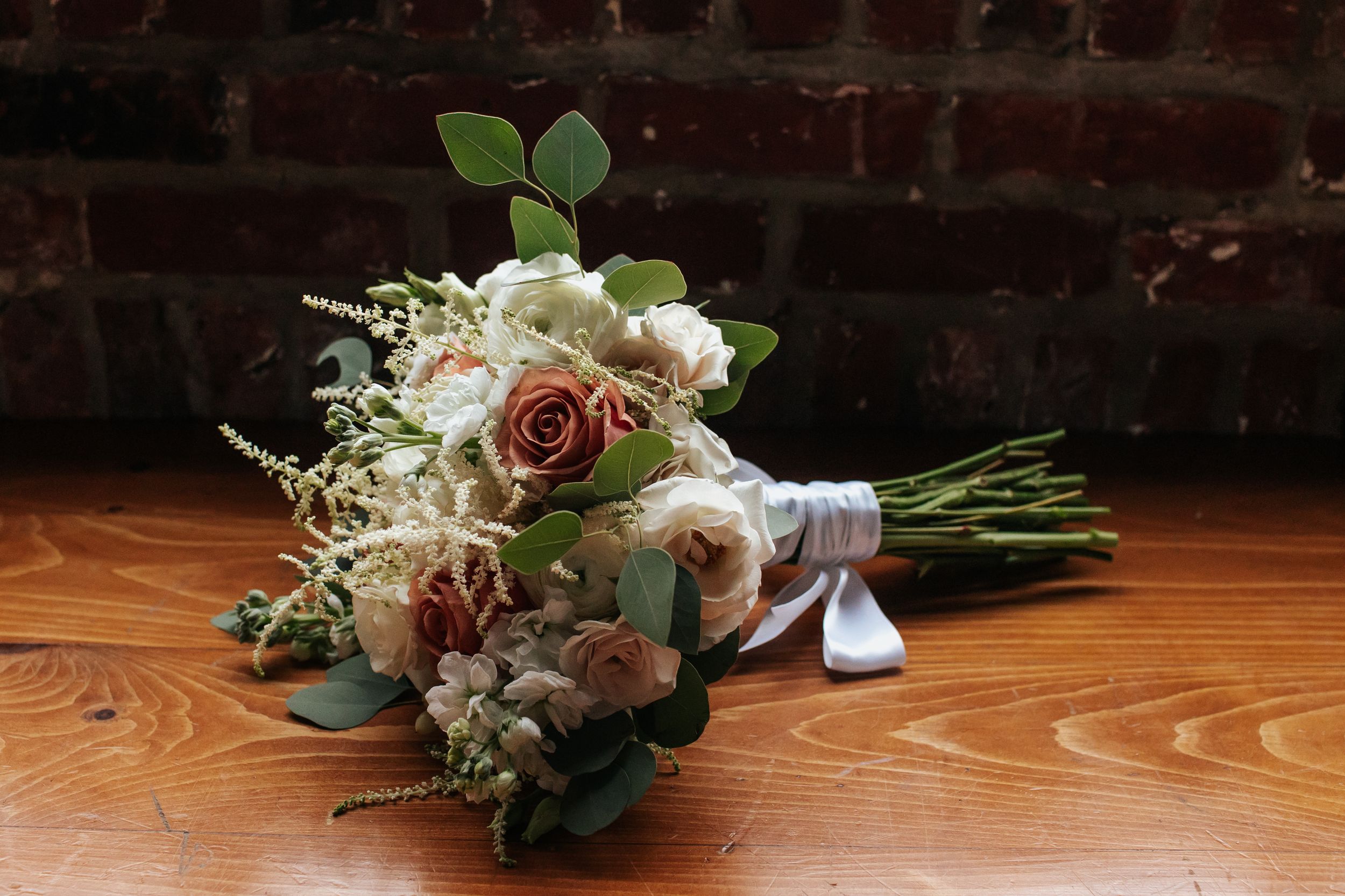 A romantic bridal bouquet with pink and white roses tied with a gray ribbon rests on a wooden surface against brick wall.