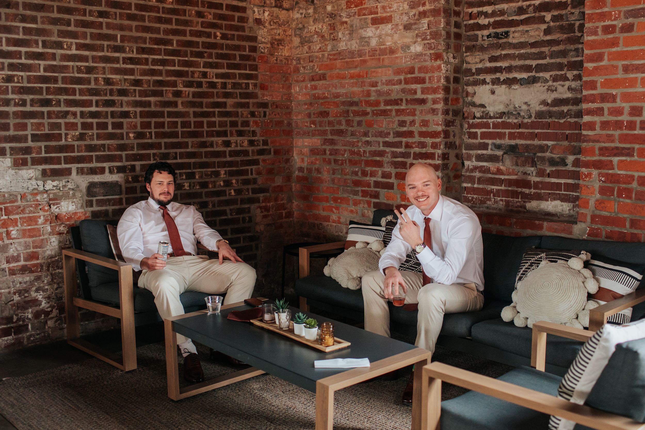 Two people in light colored attire relax on wooden chairs against a rustic brick wall during a casual gathering.