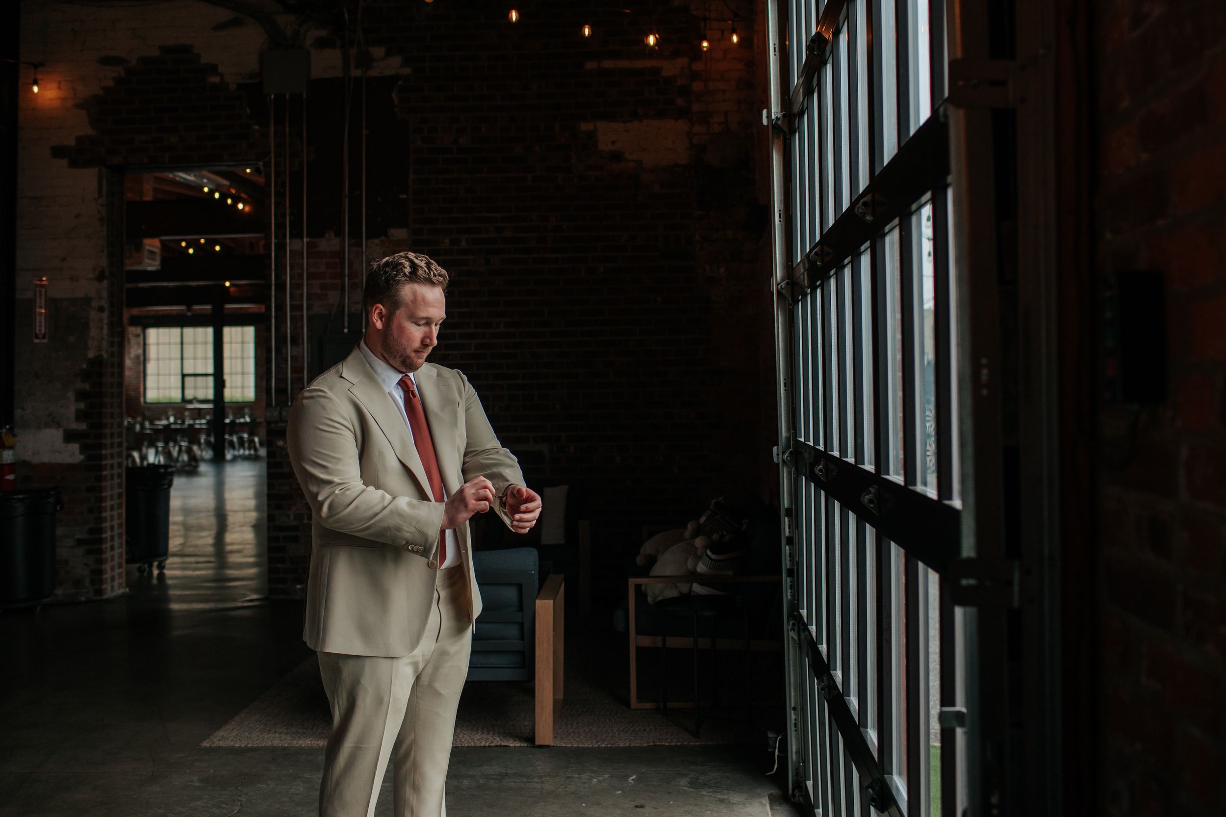 A person wearing a light colored suit stands by large windows in an industrial style room.