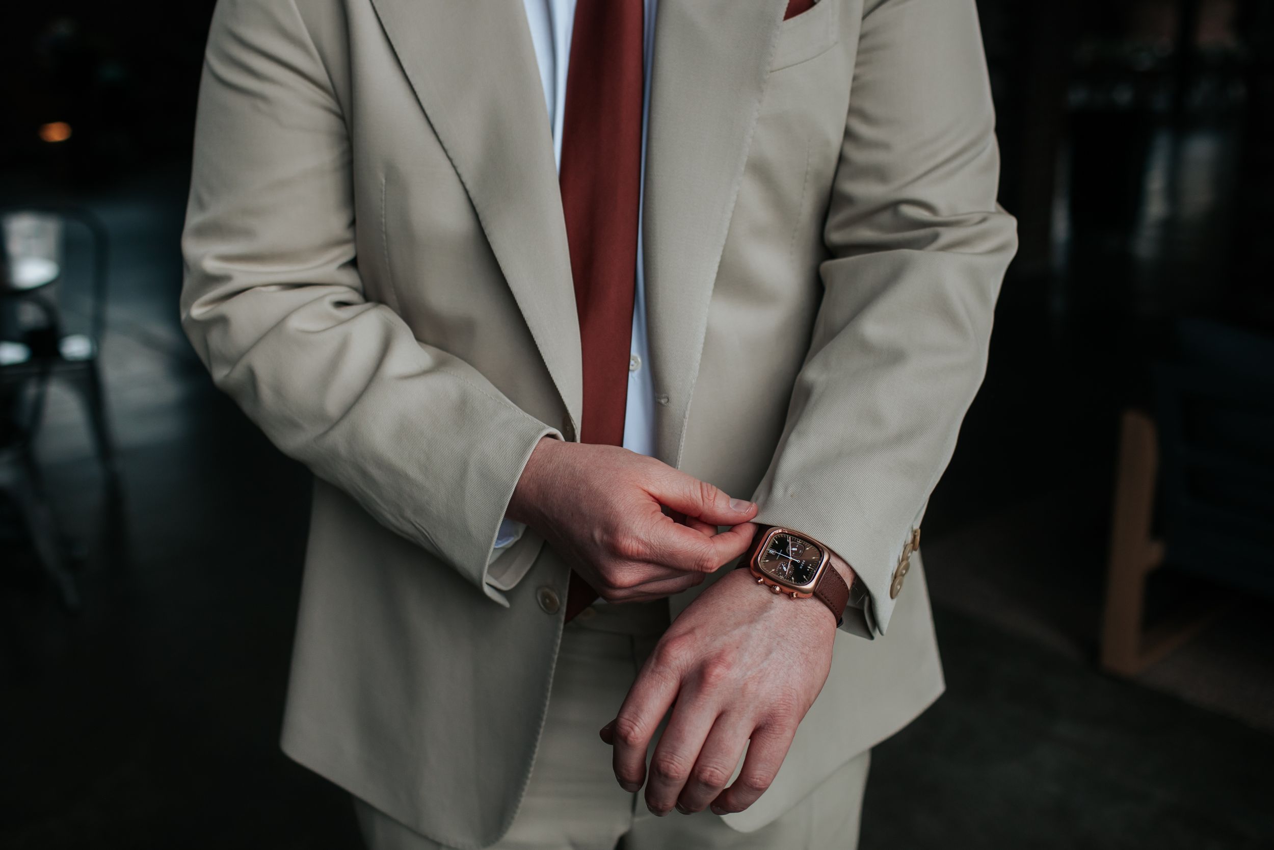 A person wearing a beige suit jacket and burgundy tie stands in a dimly lit setting.