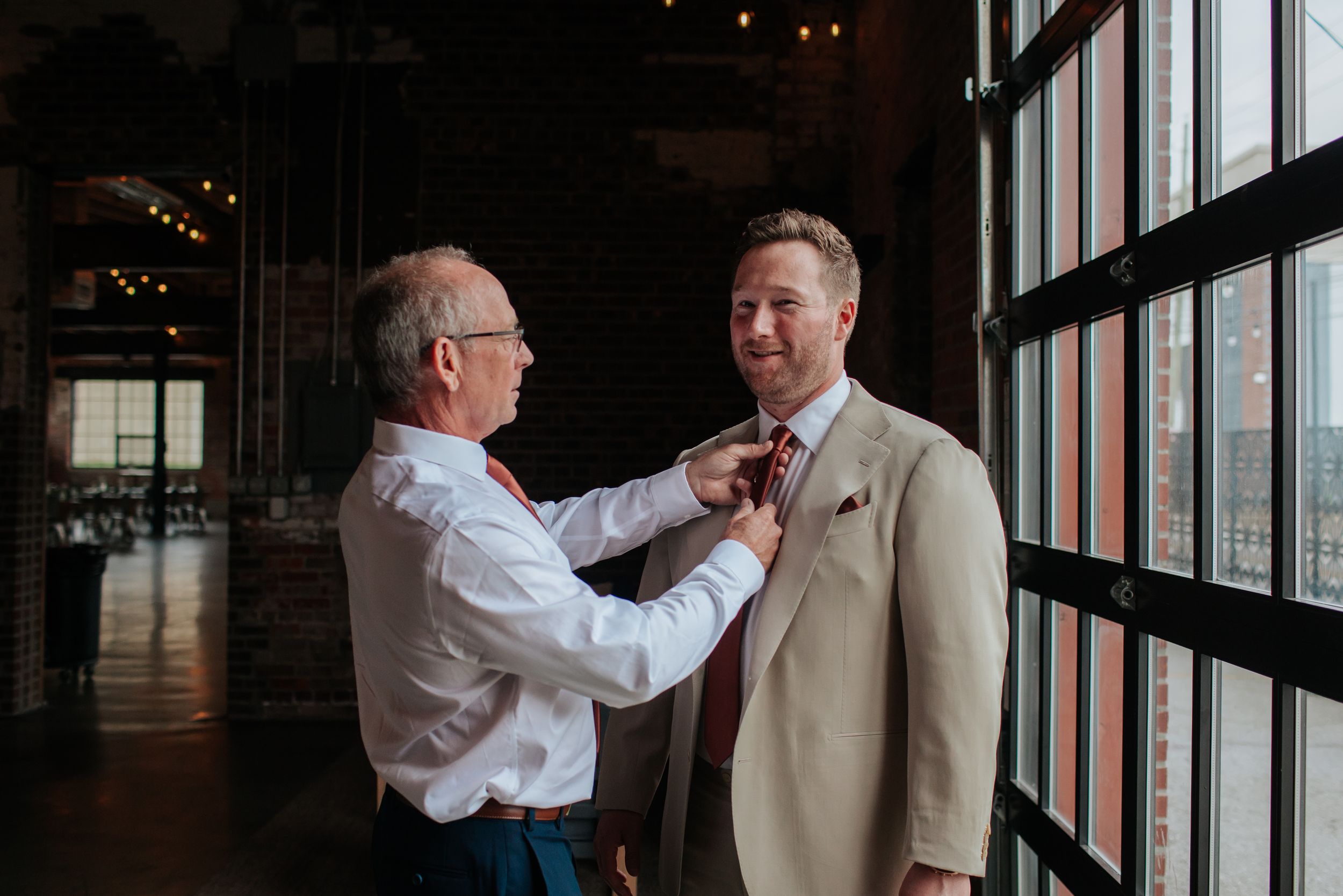 Two men in formal attire help each other with their bow ties while getting ready near large windows.