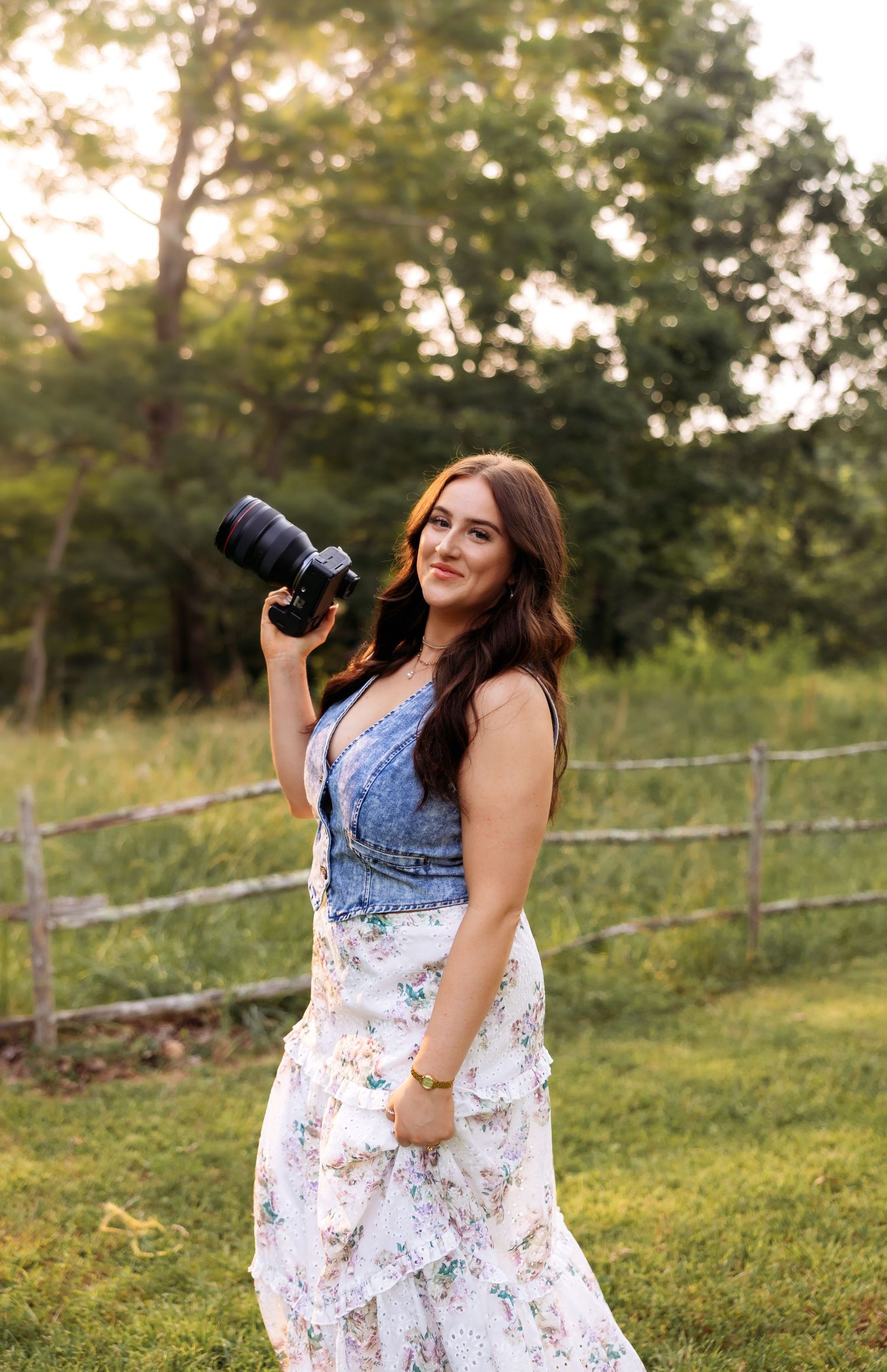 A person in a floral dress poses with camera equipment in a sunlit meadow.