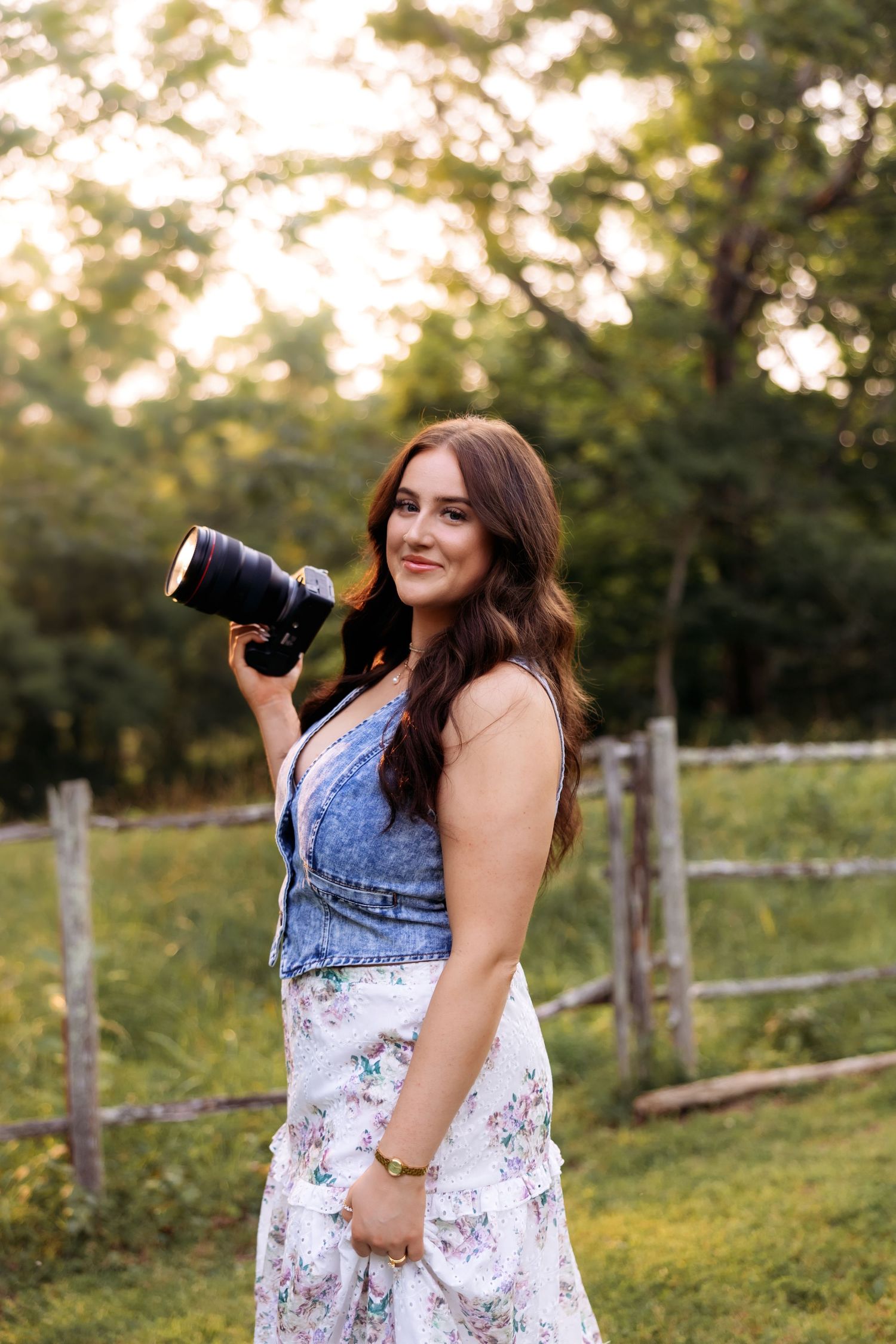 A photographer in a denim top and floral skirt holds a camera in a sunny outdoor setting.