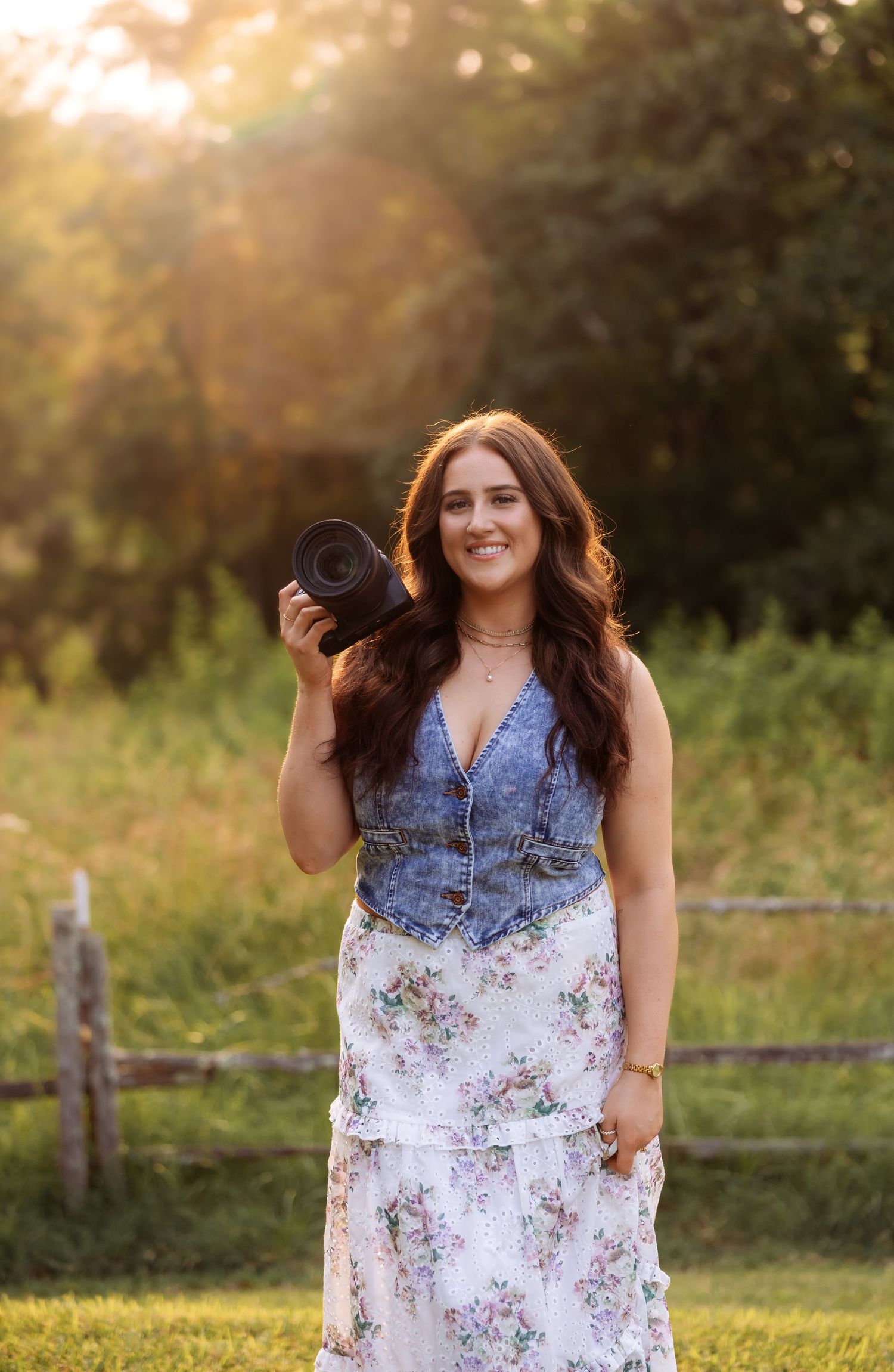 A photographer in denim and floral print poses with camera equipment at sunset.