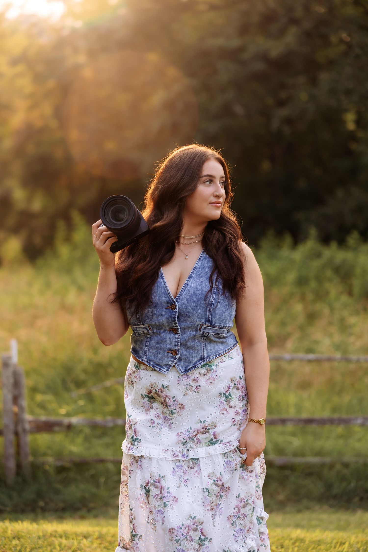 A photographer in a floral dress and denim vest poses in a sunlit field at golden hour.