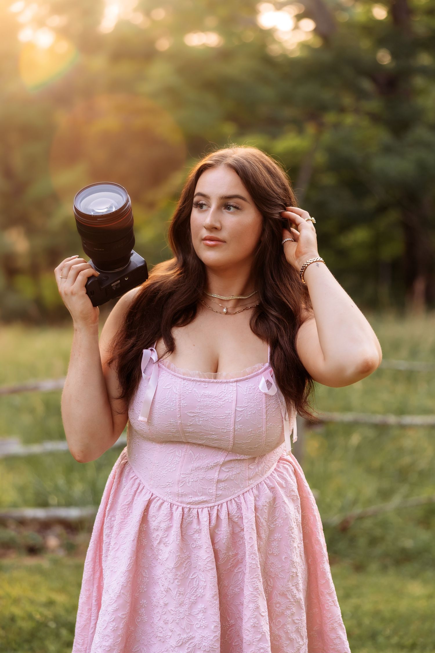 A person in a pink summer dress poses playfully in a sunlit outdoor setting with golden hour lighting.