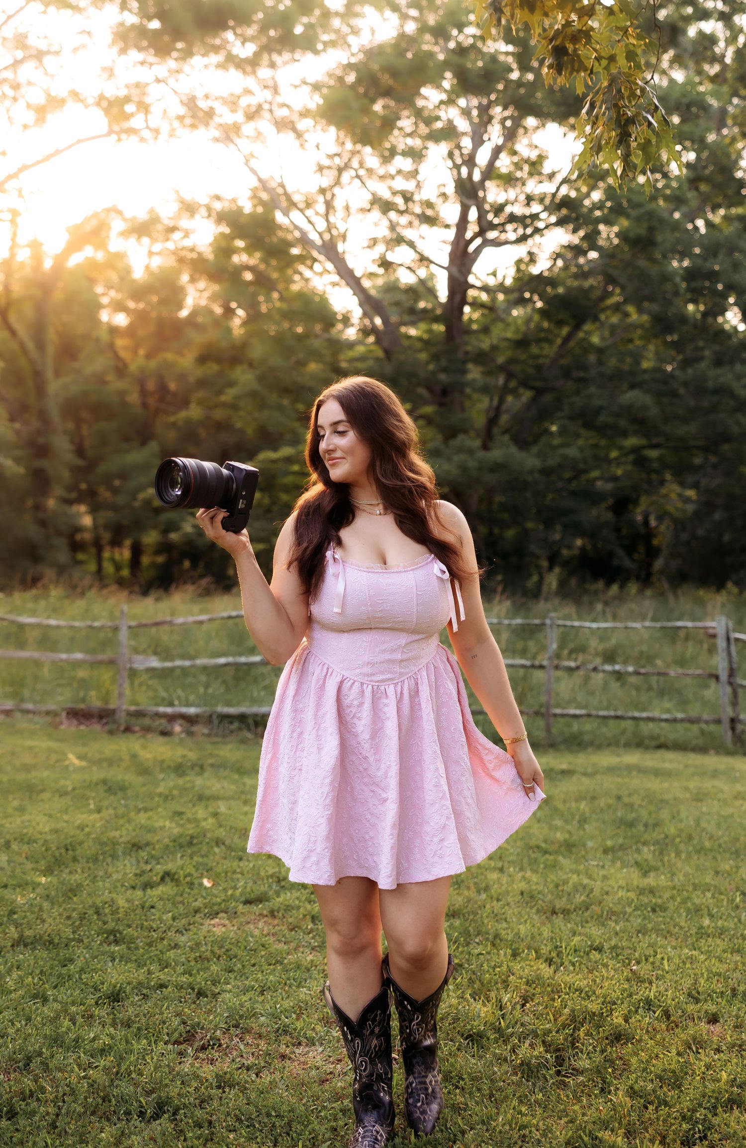 A photographer wearing a pink dress and boots twirls playfully in a grassy field at sunset.