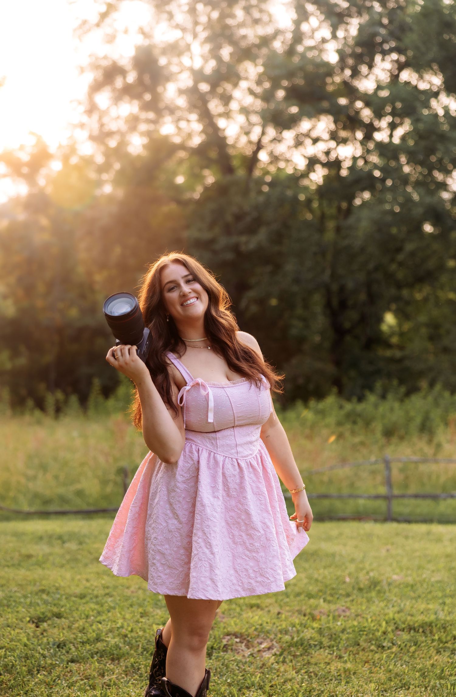 A photographer in a pink dress dances joyfully in a sunlit meadow with camera in hand.