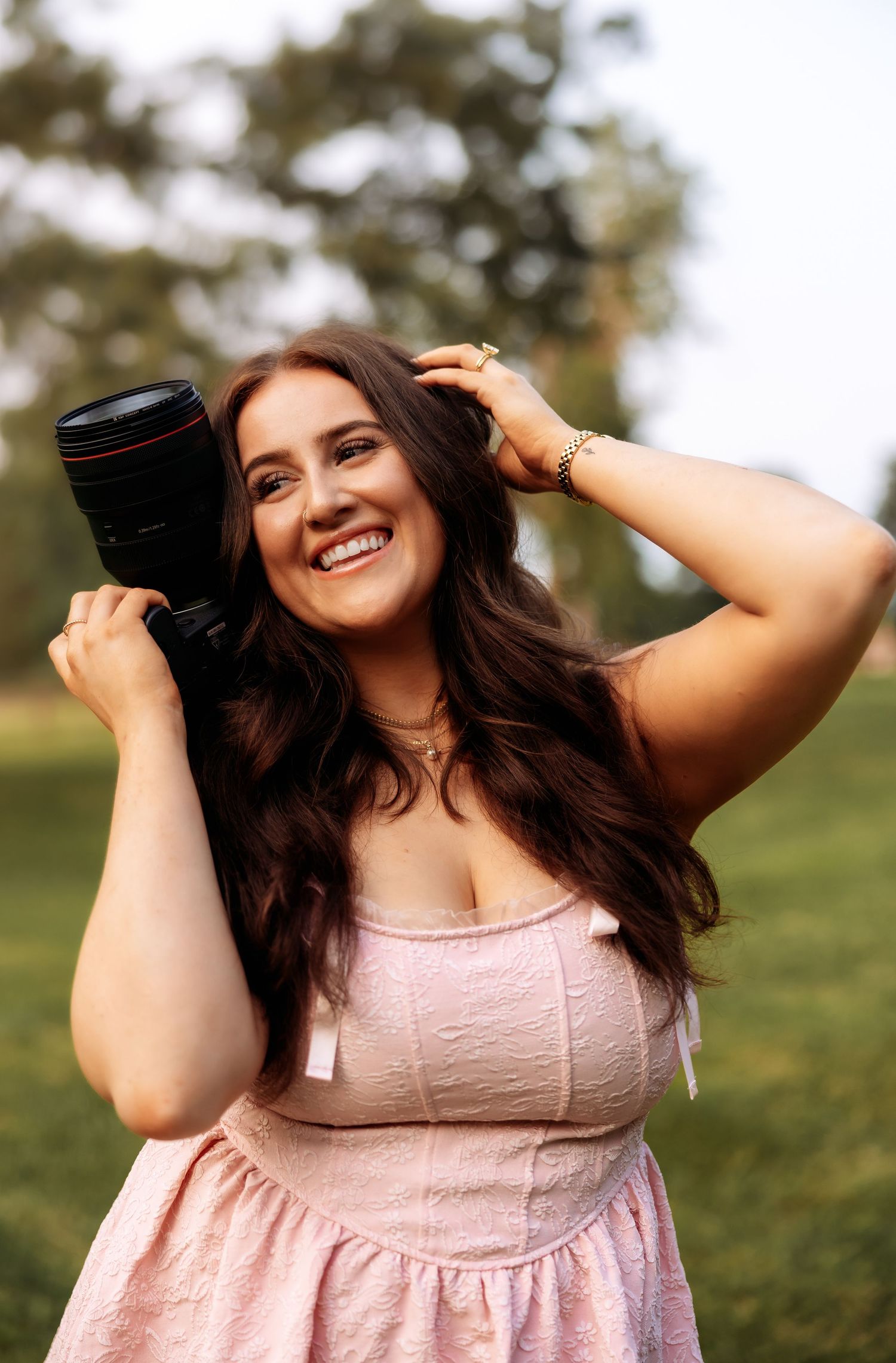 A person in a pink dress holds camera equipment while posing outdoors during golden hour.