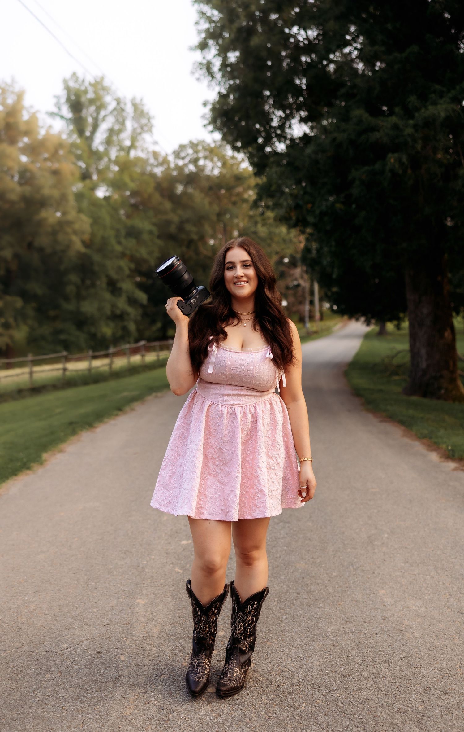A person in a pink dress and patterned boots poses on a paved path surrounded by trees.