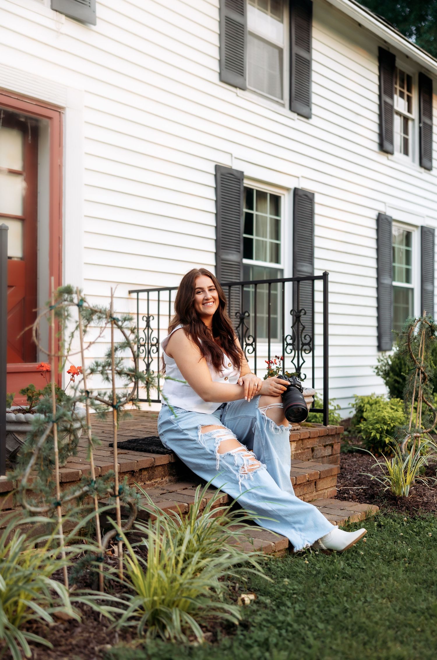 A person in casual attire sits on brick steps in front of a white house with black shutters.