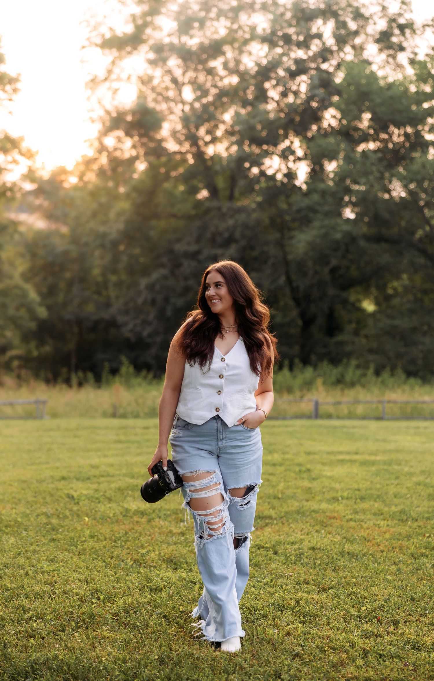 A person in ripped jeans and white top walks through a grassy field at sunset.