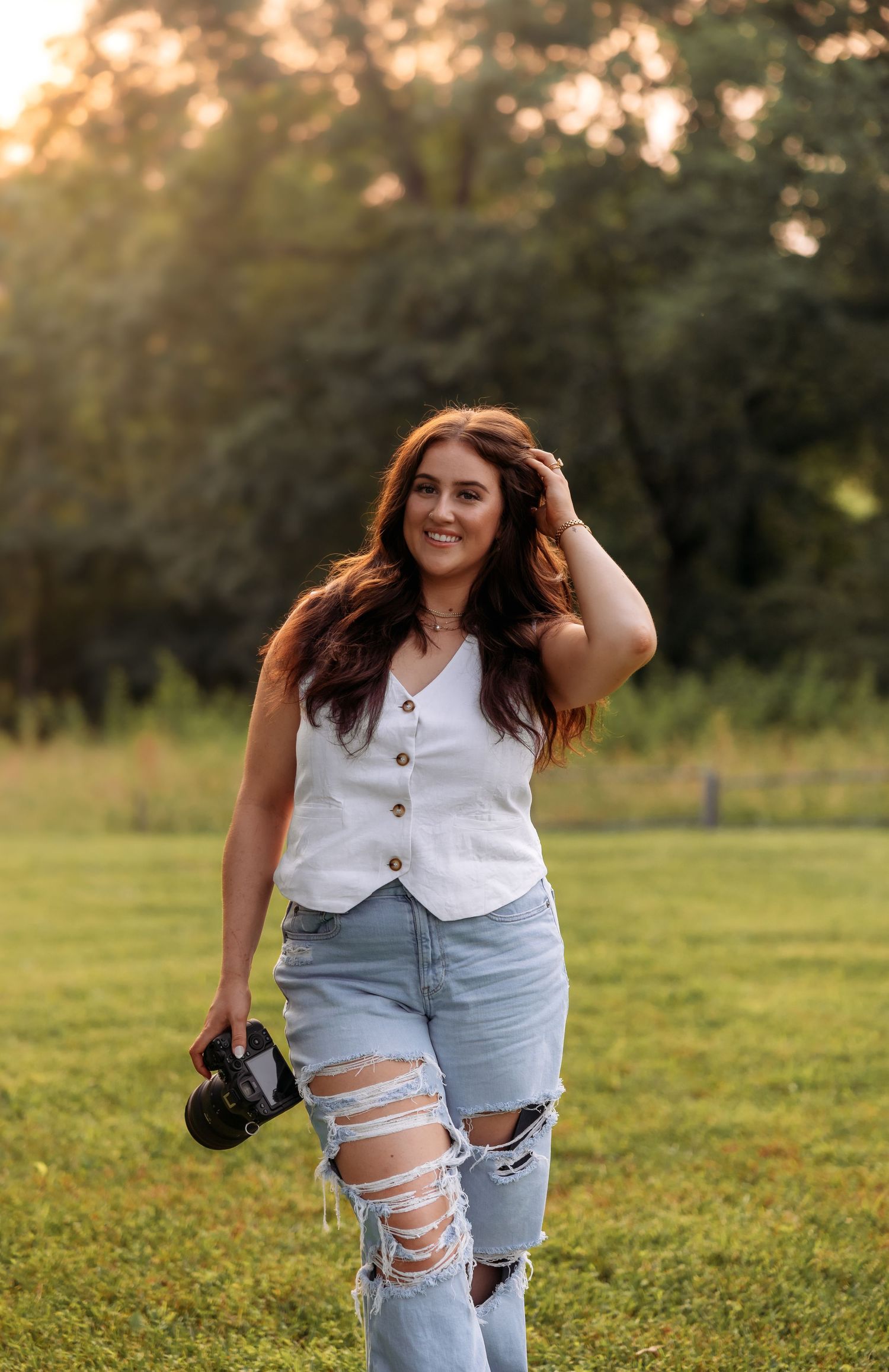 A person in distressed jeans and white vest poses with camera equipment in a sunny field.