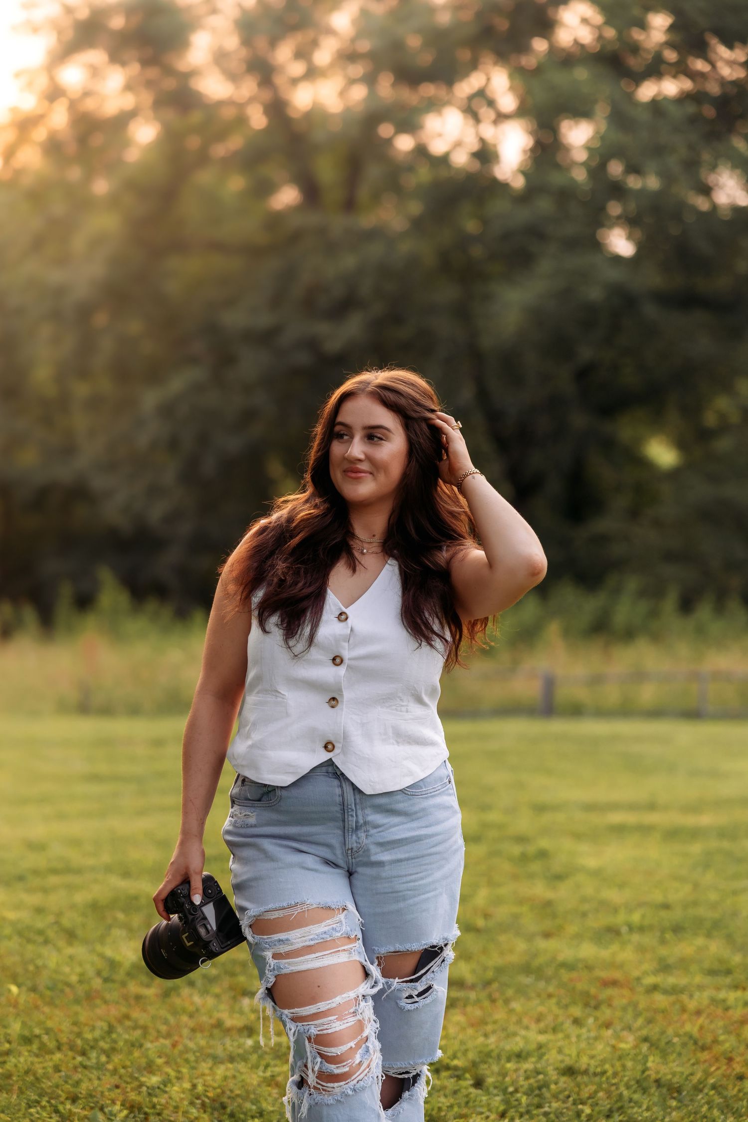 A photographer in distressed jeans and white top poses with camera in a green field.