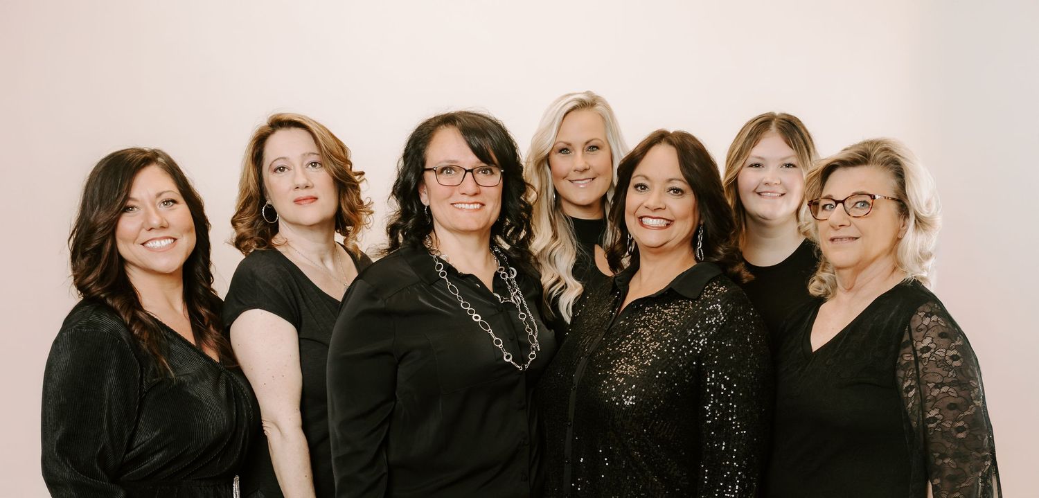 Group photo of seven women wearing black formal attire posing together against a light background.