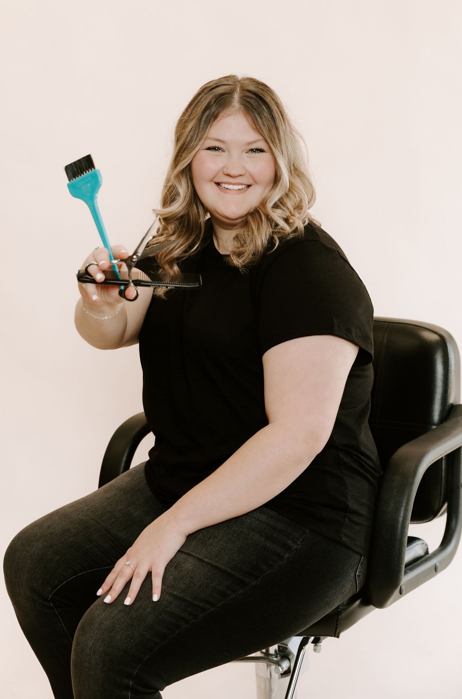 A person wearing black sits in a salon chair while holding a blue styling tool against a white background.