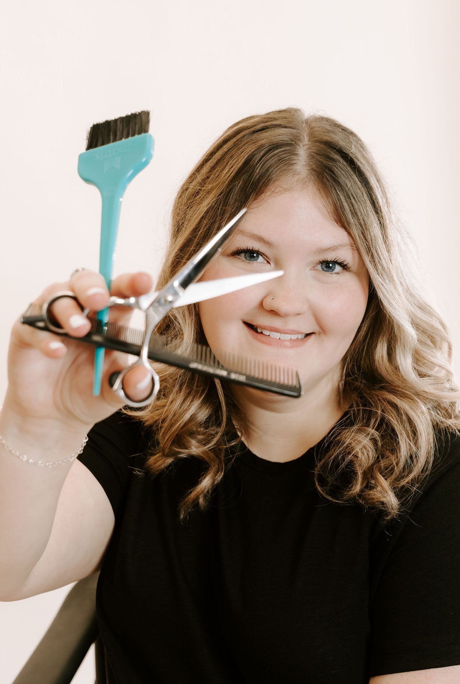 A hairstylist holds up scissors and a tinting brush while wearing a black shirt and showing wavy blonde hair.