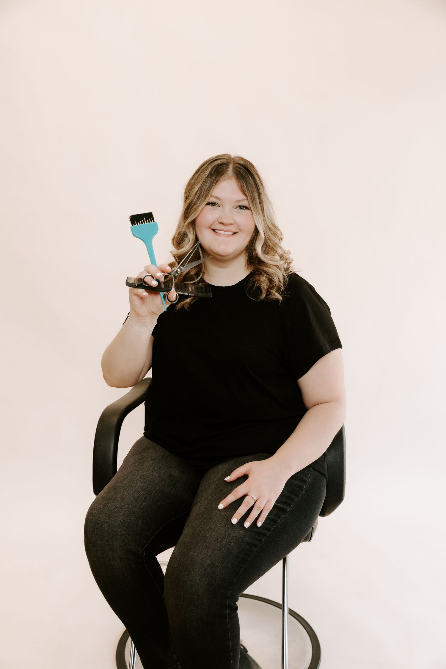 A person wearing black clothing sits in a chair holding a phone while smiling against a white background.