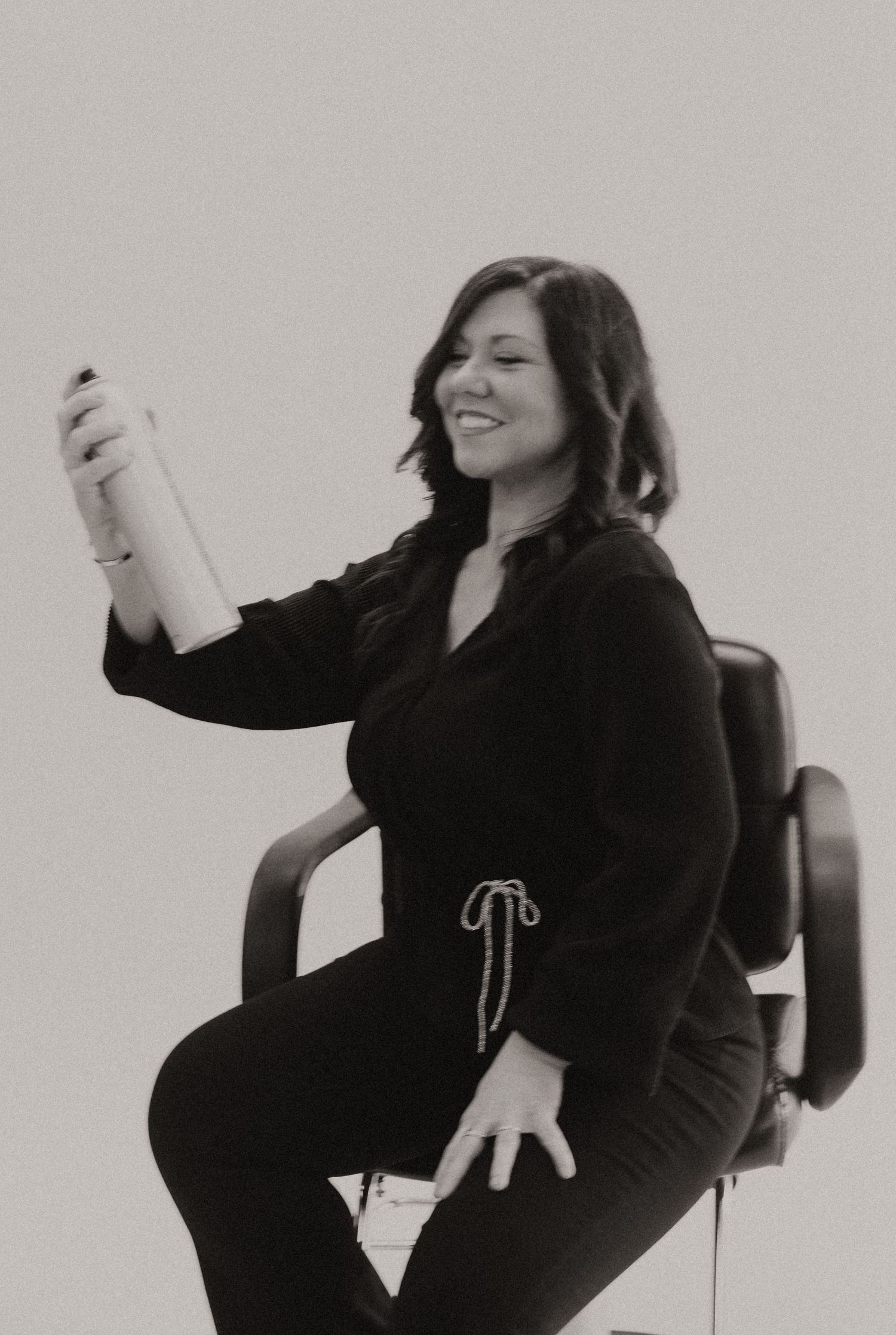 A person in a black outfit sits casually on a chair while smiling and gesturing in a black and white photograph.