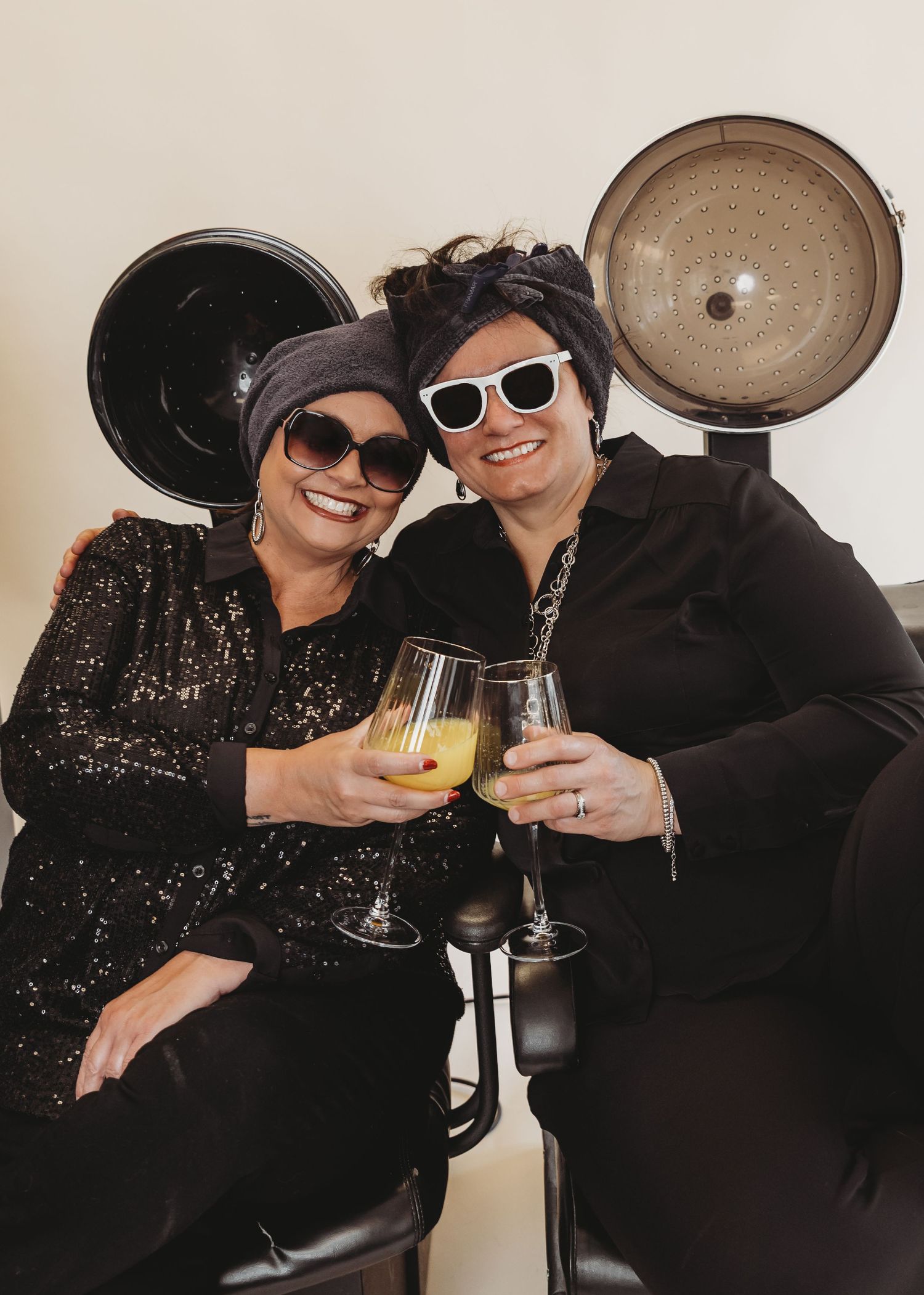 Two people in black outfits and sunglasses clink glasses in a celebratory toast.