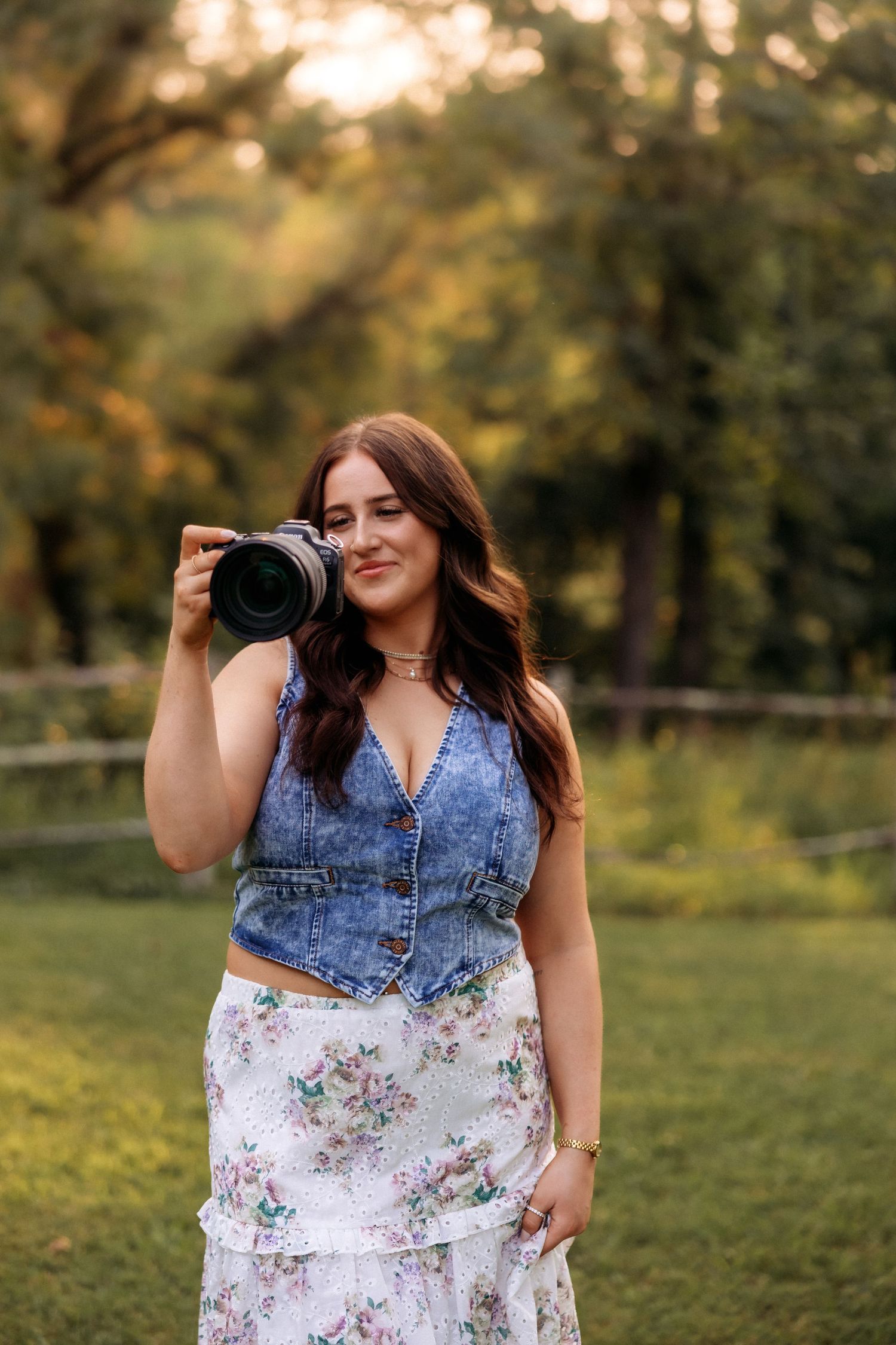 A photographer in denim vest and floral dress poses with camera in a sunlit field.