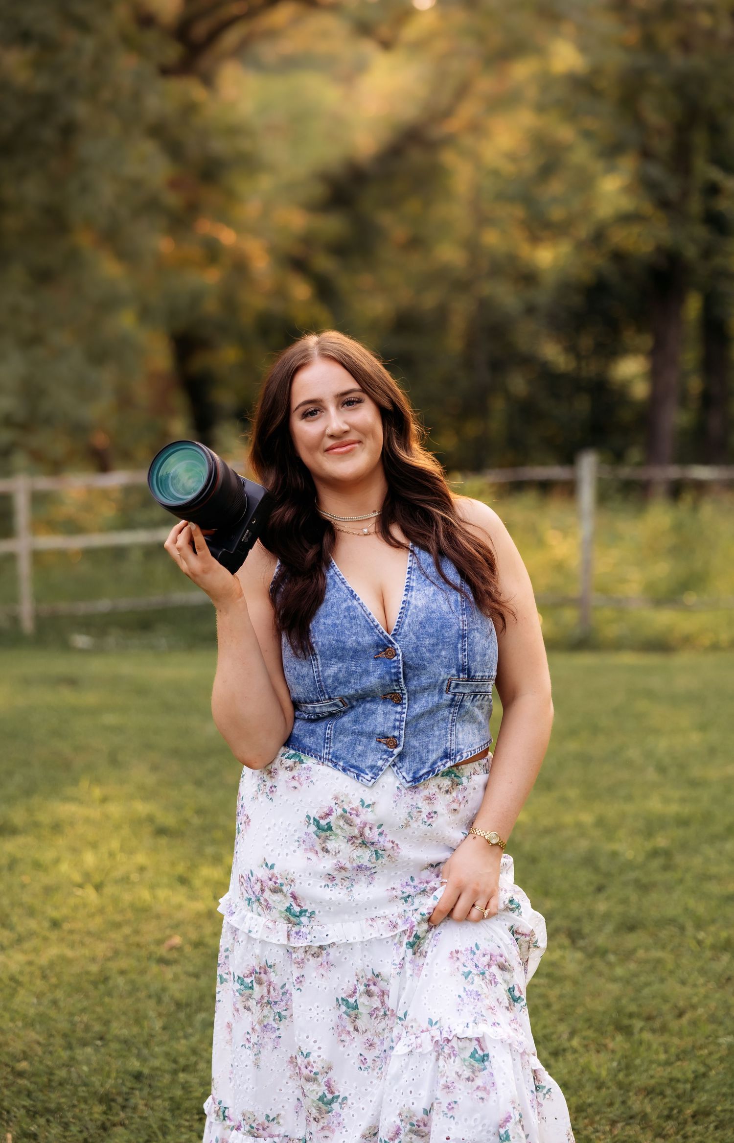 A photographer in a denim and floral dress poses with a camera in a sunny outdoor field.