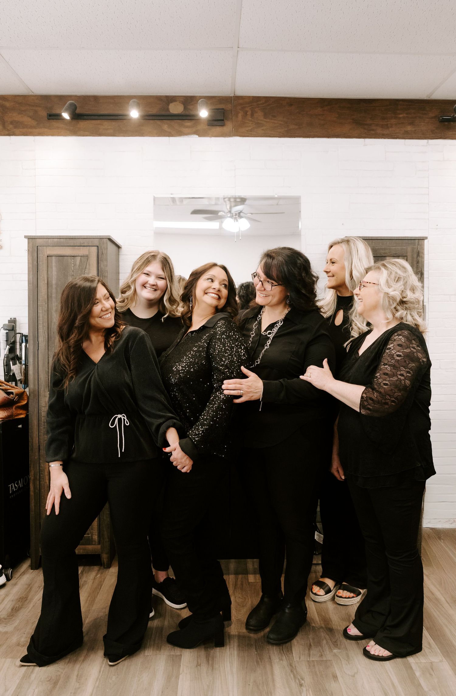 A group of women in black outfits posing together in a dance studio with white walls and wooden floors.