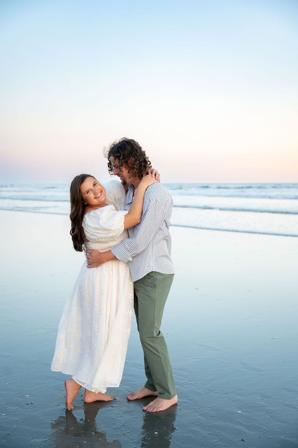 A couple dances playfully on a serene beach at sunset, the woman wearing a white dress and the man in green pants.